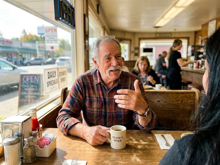 A man in his late sixties in a plaid shirt sitting at a diner table with a coffee mug, talking with his hands.