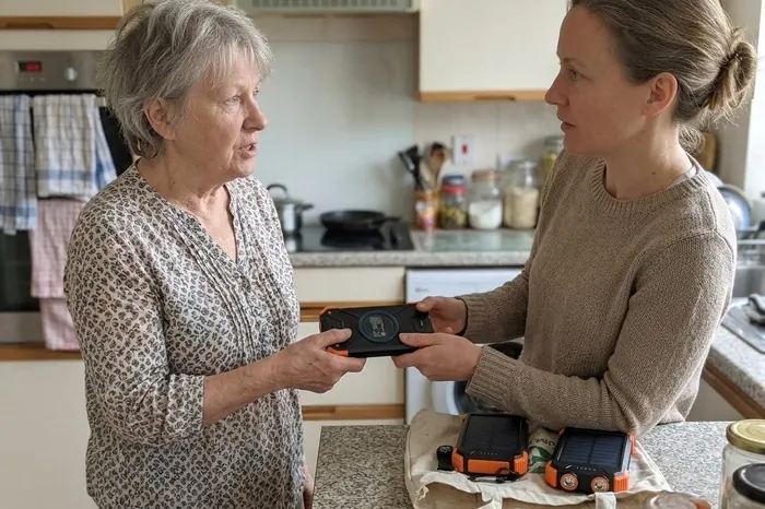An older woman handing a solar power bank to her adult daughter in a kitchen, with two more on the counter